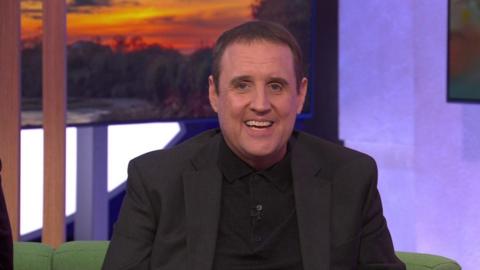 Peter Kay sits on a green sofa, he wears a black knitted polo shirt under a black jacket. He has short dark hair and smiles widely at the camera