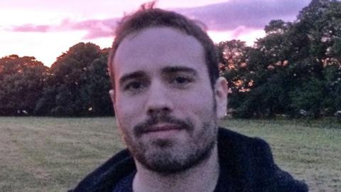 A man stood in a field. He is wearing a hoodie and smiling.