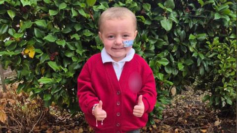 A young girl with short blonde hair and a tube taped into her nose. She is wearing a red school cardigan and smiling with both thumbs up. She is standing in front of a hedge.