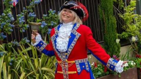 Kila Redfearn smiling and holding her bell. She is in full red white and blue town crier regalia with a frilly white blouse and a long red jacket with blue and gold decoration and a black tri-cornered hat with a red feather. She has blond hair and is smiling in a garden