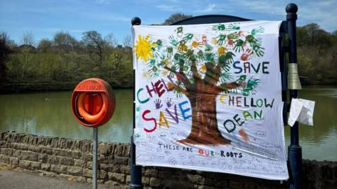 A poster which reads 'save Chellow Dene' erected near a reservoir.