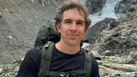Image shows Alec Luhn carrying a backpack,  with mountains in the backdrop