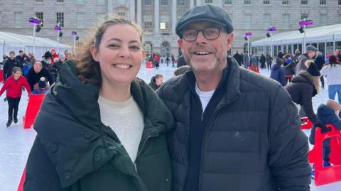 Alice, smiling and wearing a warm green jacket alongside her father Nick Robinson, who is wearing glasses and a flat cap hat and dark jacket