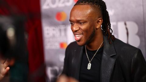 KSI is wearing a black blazer and t-shirt with a silver necklace.
