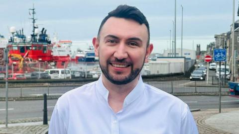 A man with dark hair and a dark beard smiles at the camera while standing in front of a harbour