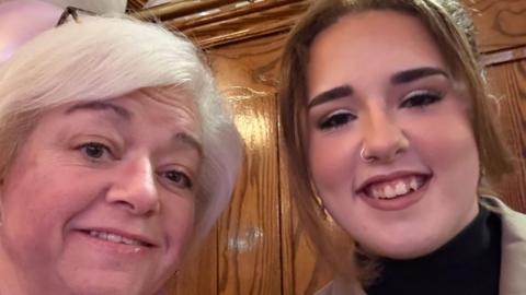 Andrea with short white hair. She is smiling. Ibbie has brown hair tied back and a nose ring. She has a beige jacket and black top. She is smiling.