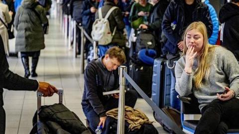 Travelers queue with their luggage at Schiphol Airport. One woman is sitting with her phone in her hand, while a man behind her is kneeling next to his bag while reading a book.