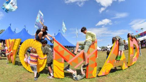 WOMAD sign in orange, yellow, brown and pink. Children are climbing on the letters and there is a blue big top tent in the background.