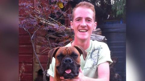 Sam Boulton wearing a green t shirt smiling with his hand on a brown dog, which has its tongue out.
