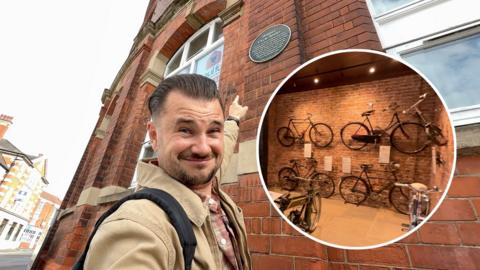 The image shows Josh Gorroño Chapman pointing at a commemorative plaque on the exterior of a historic red-brick building with arched windows. An inset reveals an indoor display of vintage bicycles mounted on a brick wall.