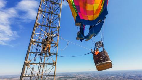 A metal tower with people on it using ropes tied to a hot air balloon with people in it.