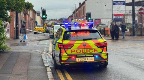 A parked police car in front of a closed Maynard Road in Leicester. More emergency service vehicles can be seen in the background with police tape across the road. Passersby have stopped on the street corner to observe what has happened. 