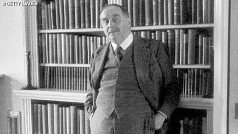 Black and white image of AG Wells standing in front of a bookshelf, with his hands in his pockets, looking at the camera, wearing a three piece suit.