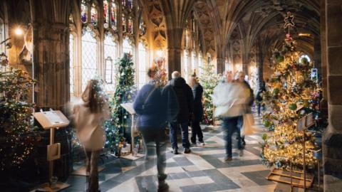 A time lapse image showing blurred people walking through a hallway at Worcester Cathedral. The hallway has Christmas trees on either side of the walkway with large windows to the left with light coming through.