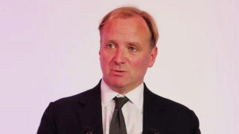 GMB boss Gary Smith, a man wearing a white shirt and dark-coloured tie with a black suit jacket. He is stood against a light purple backdrop.