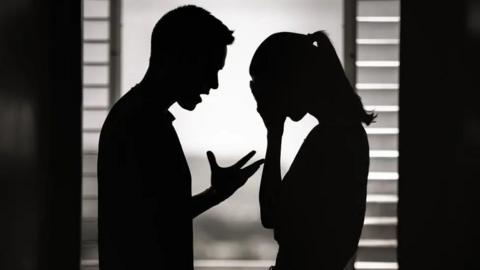 File picture of the silhouettes of a man and a woman apparently arguing - the man has the fingers of his right hand stretched out and he is looking at the woman standing close to him, who has her head in her hands. There are closed blinds behind them.