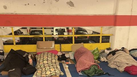 People who took part in the event are sleeping on the car park floor. The participants are tucked up in sleeping bags and blankets. They are lying side by side. A strip of cardboard separates them from the cold hard floor. Cups, water bottles and plastic bags have been placed around them. 