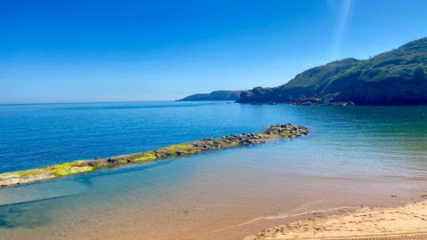 La Grève de Lecq in Jersey. There is blue clear water. The sky is a lighy blue. In the foreground of the picture is sant. In the background to the right is a green hill. 