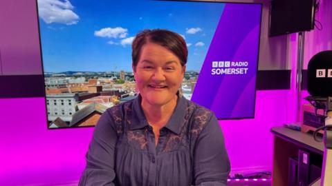 Emma Britton in the BBC Radio Somerset studio. She has short dark hair, is wearing a blue top in a purple radio studio with a screen and a microphone.