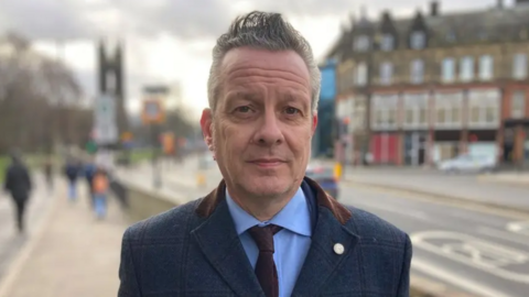Nick Kemp, with short grey hair, is standing on a street. He is wearing a blue suit and is looking at the camera, expressionless.