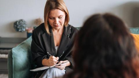 A woman sits in a sofa and makes notes on a clipboard, while a woman (seen from behind) sits opposite her. They are in a room with grey walls and the sofa is green.