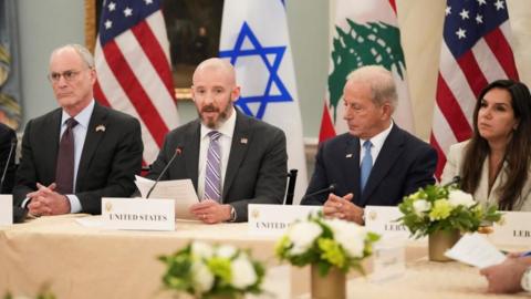 Three men and one woman, all dressed in suits, sit at a long table in front of the US, Israeli and Lebanese flags.
