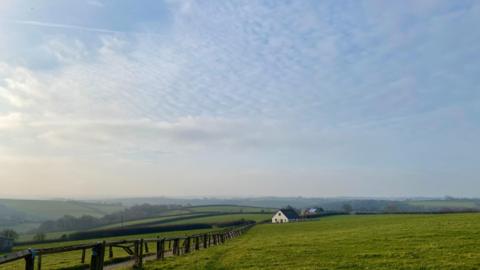 Rural countryside with hills and hedgerows in distance, a wooden railing to the left and a white house in the background, under a blue sky with wispy cloud