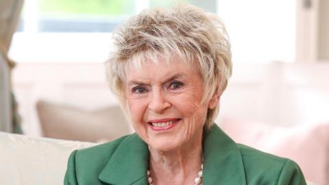 Gloria Hunniford is smiling toward the camera. She has short blonde hair and is wearing a green blazer and white pearls.