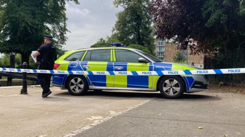 A police car is parked across a footpath near a river. A police officer is checking paperwork with his back to the camera. Blue and white police scenes of crime tape has been placed in front of him attached to a metal bollard to the left and iron fencing to the right.
A block of flats is visible in the background on the other side of the river.