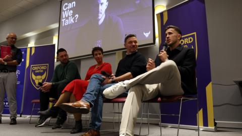 Des Buckingham, Jess Wilshire, Luke Beauchamp and Will Vaulks are all sat down on chairs. Behind them is a black screen with the white writing 'Can We Talk?'.