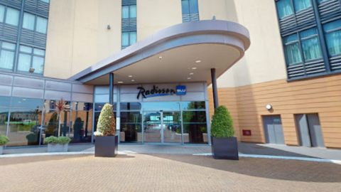 A picture of the front of the Radisson blu in Jersey. It is an all brick building with glass windows and plants in front of it. There is a large glass main entrance in the middle of the image.