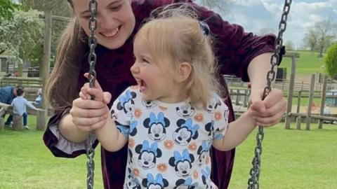 Isabella smiling on a swing with her mother smiling and standing behind her. Isabella has a T-shirt on with Minnie Mouse's face on it.