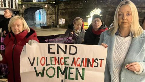 Two women, one in a red coat and another in a blue coat, both hold up a sign which reads: 'End Violence Against Women + Girls' at the rally in Guildhall Square.