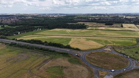 Aerial view of the proposed site. There is a large expanse of grassland to the side of a roundabout, with housing in the distance.
