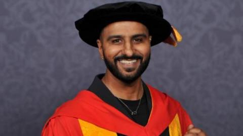 Harpreet Singh Jandu wearing a red and orange gown, holding a graduation scroll