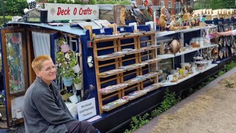 A woman sits at the end of a canal boat will shelves hanging alongside it selling various items. There is a sign on top that says "Deck O Crafts".