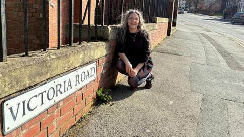Councillor Marina Strinkovsky leans down next to a road sign that reads "Victoria Road".
