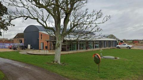 The exterior of a large farm building. Grass and a large tree sits in front of it. A road leads up to the building and gravel where cars are parked. The sky is overcast.