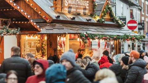 Crowds of people on a shopping street in front of two market stalls covered in festive lights. There are shop buildings in the background