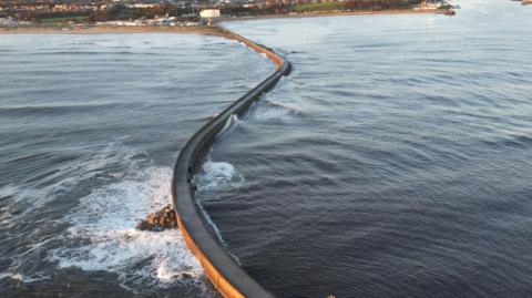 A pier with a road on it stretches out into the blue ocean. There are rocks dotting the border between the pier and the water. The town of South Shields can be seen in the background.