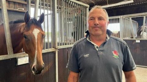 A man in a grey polo shirt stands facing the camera to the right of the image. He is stood in a stables. To his left, a brown horse is sticking its head out of its stable and also looking at the camera. There are more horses behind him.
