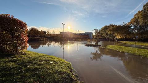 A flooded outdoor area with water covering a road and grassy sections, reflecting trees and lampposts in the sunlight. The sun is low in the sky, casting a warm glow over autumn-colored foliage on the left and trees on the right. A building is visible in the background under a partly cloudy blue sky.