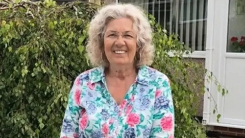 Valerie Kneale, with shoulder-length grey curly hair, wears a pink, lilac and green floral blouse as she is photographed in a garden. She is standing in front of a pebble-dashed house with a white plastic door and windows.