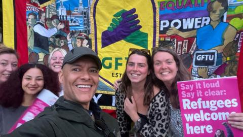 Clive Lewis MP, wearing a dark baseball cap, in a selfie surrounded by five unknown women, one of whom holds a poster which says "Say it loud, say it clear: Refugees welcome here". They are standing in front of a fabric banner which shows two clasping hands, GFTU, and the words "equality" and "power"