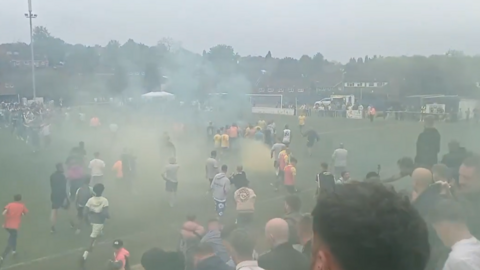 An image taken from the stands of a football ground, showing dozens of people on the pitch. The view is obscured by clouds of green and yellow smoke drifting across the pitch.