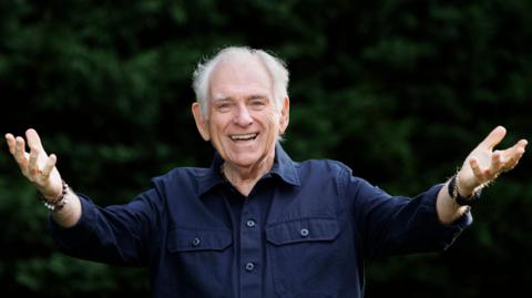 Hugo Duncan smiling at the camera with his arms outstretched. He is wearing a blue shirt. Behind him there are some bushes