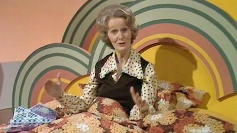 A woman TV presenter sat under a duvet in a double bed in the studio.