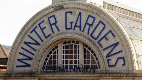 Semi circular tiled sign saying Winter Gardens in blue on cream on the outside of the building