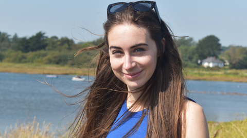 Bethany Handley is a white woman in her mid 20s. She has long light brown hair with sunglasses on top of her head in a head and shoulders shot. She wears a blue vest and is smiling at the camera. She is outdoors in front of a lake which is out of focus in the background surrounded by grass and trees.