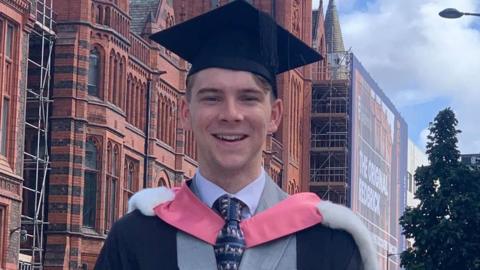 Lucas Martin, a young man with dark hair smiles brightly. He has a graduation cap and gown and wears a grey suit as he stands in front of an old red brick building.